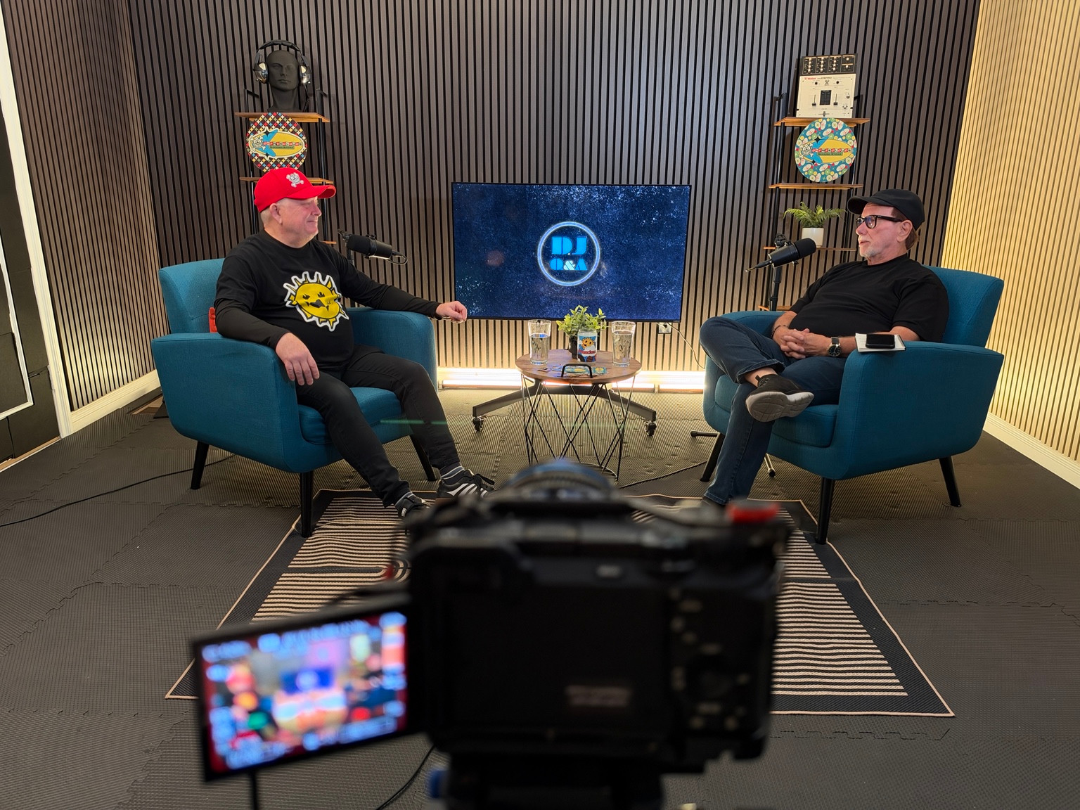A camera in the foreground of a two-person studio interview.